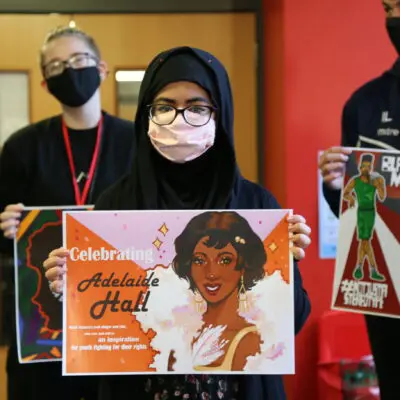 a young woman holds her graphic designed poster with more students stood behind her with their efforts