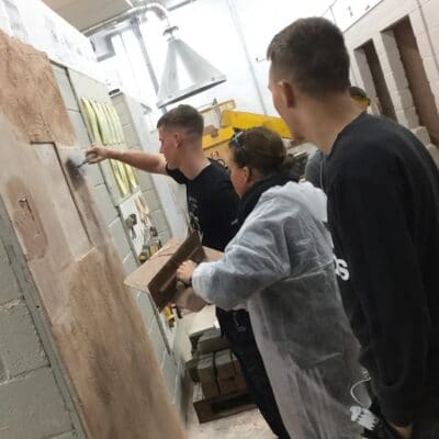 A Maths tutor is directed by two students in front of a wall