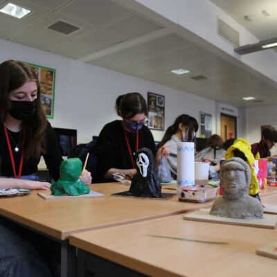 A group of students sit at a desk in masks painting models.