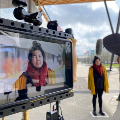 A woman stands being interviewed with the camera mirroring this in the far left of the picture.