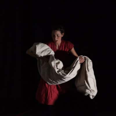 Joanne in a play stood against a black background carrying a sheet in her arms.