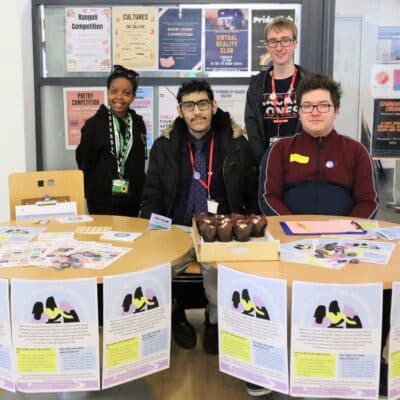 2 students sat in front of two tables and two students standing beside them