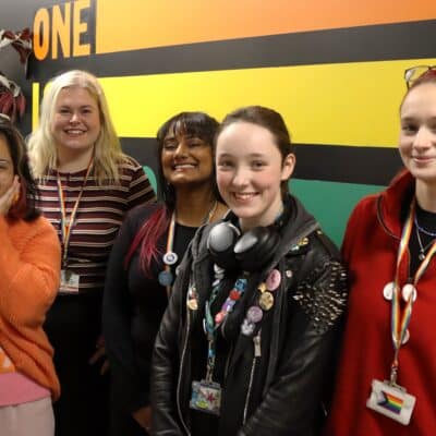 5 people standing in front of flag smiling at College prestigious Rainbow Flag Award